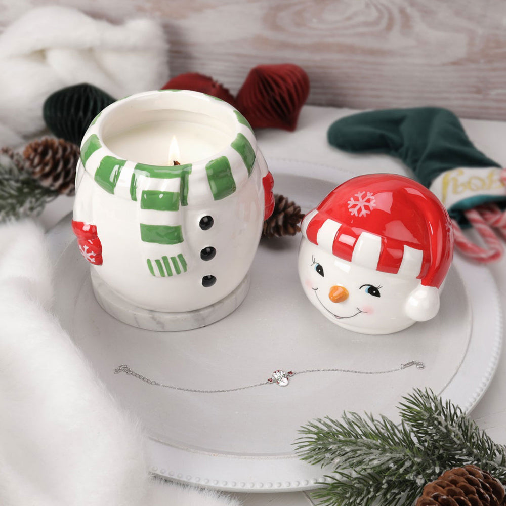 Frosty Jewel Candle - weihnachtliche Schneemann-Duftkerze mit grünem Schal und roter Mütze, brennend auf Tablett mit Armband-Überraschung, Deko und Tannenzweigen.