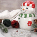 Frosty Jewel Candle - festliche Schneemann-Duftkerze mit roter Mütze und grünem Schal, dekoriert mit Weihnachtskugeln, Tannenzweigen und Schmuck-Armband als Überraschung.